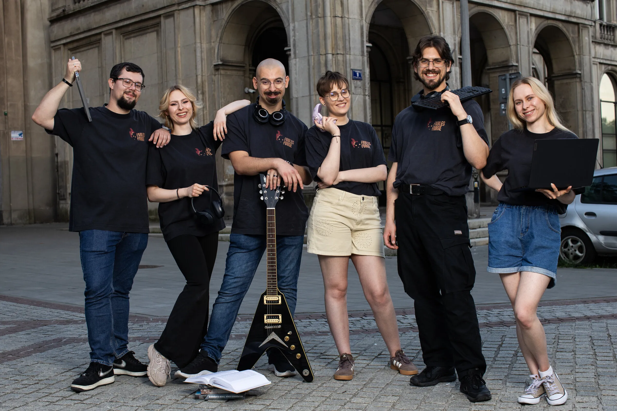 The Fables Forge team in studio T-shirts, posing with a guitar, headphones, a keyboard and a laptop in front of an arcaded building